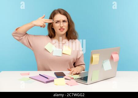 Depresso dipendente donna overworked in occhiali nerd seduto sul posto di lavoro, tutti coperti di note appiccicose, sparando con finger gun suicide g Foto Stock