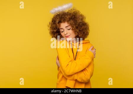 Mi amo! Ritratto di bella donna angelica con capelli ricci, con san nimbus abbracciandosi e sorridendo di piacere, sentendo compiacenza. In Foto Stock