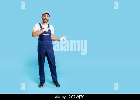 Handyman sorpreso di lunghezza intera in uniforme che punta allo spazio di copia, l'area di pubblicità per la presentazione di idea, guardando scioccato alla macchina fotografica. Professione di ser Foto Stock