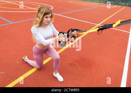 Bella giovane bionda caucasica donna in abiti sportivi rosa, allenandosi con allenatore di sospensione all'aperto presso il campo da tennis rivestito rosso. Fitness Foto Stock