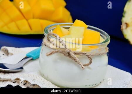 Vaso in vetro con cubetti di yogurt e ananas e mango su sfondo di legno blu Foto Stock