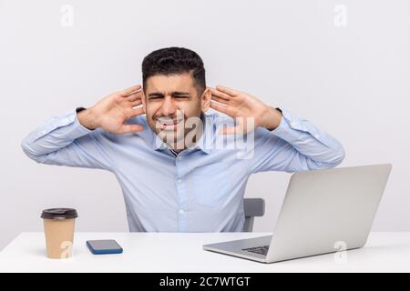 Uomo confuso che lavora sul posto di lavoro con il computer portatile, tenendo le mani vicino alle orecchie cercando di sentire meglio, difficile da ascoltare in un ufficio rumoroso. Indoo Foto Stock