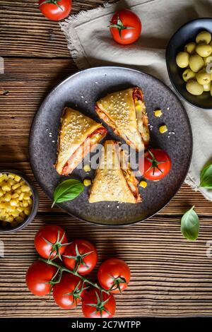 Deliziosi panini triangolari per pizza con pasta sfoglia ripieni di salsa di pomodoro, prosciutto, formaggio, mais, olive e cosparsi di semi di sesamo Foto Stock