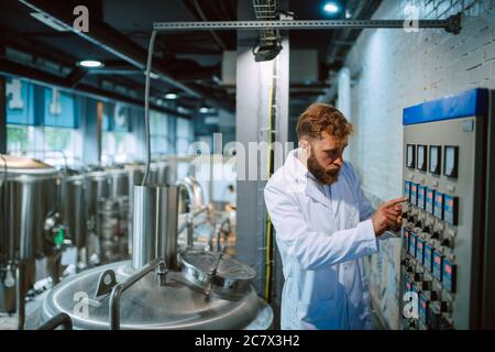 Operatore di quadri di controllo macchina in impianto di produzione. Specialista tecnico caucasico in tuta bianca che lavora in fabbrica di alimenti e bevande Foto Stock