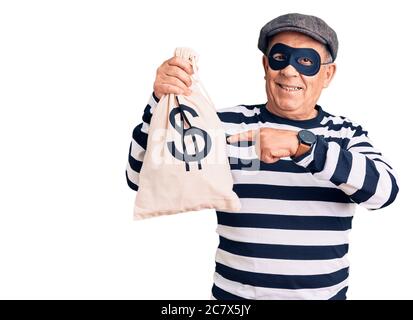 Uomo anziano bello che indossa maschera di scasso tenendo borsa di denaro sorridente felice di puntare con mano e dito Foto Stock