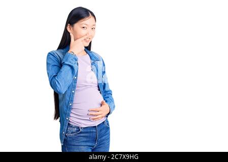 Giovane bella donna cinese incinta aspettandosi il bambino che punta con la mano dito a viso e naso, sorridente allegro. Concetto di bellezza Foto Stock