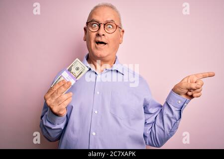 Uomo di mezza età che tiene mazzo di banconote di dollari su sfondo rosa isolato molto felice indicando con mano e dito a lato Foto Stock