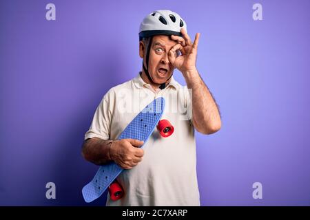 Uomo di mezza età che indossa casco di sicurezza che tiene skateboard su sfondo viola con faccia felice sorridente facendo segno ok con mano su occhio che guarda Foto Stock