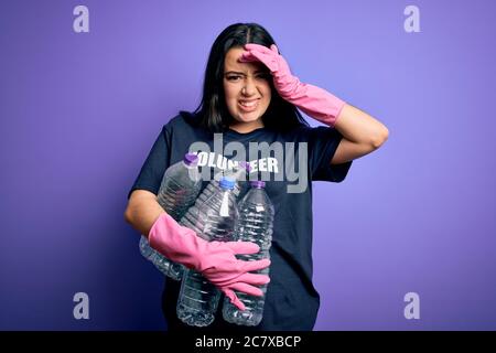 Giovane donna bruna che indossa t-shirt volontaria picking bottiglie di plastica su sfondo viola stressato con mano sulla testa, scioccato con vergogna e surpr Foto Stock