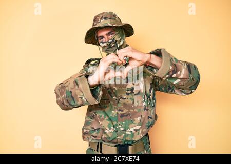 Giovane bell'uomo che indossa uniforme armata camouflage e balaclava sorridente nell'amore mostrando il simbolo del cuore e la forma con le mani. Concetto romantico. Foto Stock