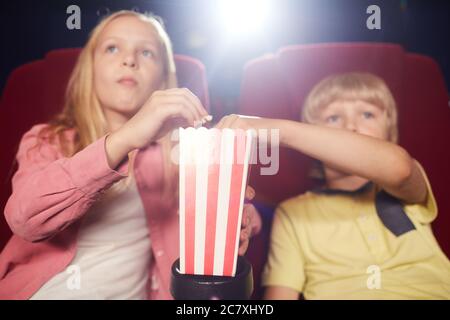 Primo piano di due bambini biondi che condividono la tazza di popcorn mentre guardano i cartoni animati nel cinema, concentrati su primo piano, copia spazio Foto Stock