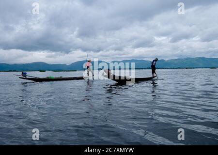 Due pescatori in piedi su piccola barca nel lago Inle increspato. Utilizzo di una barca da gamba a fila. Cielo bianco nuvoloso con montagne Foto Stock