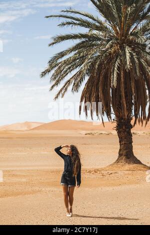 Donna che cammina nel bellissimo deserto con dune di sabbia e una sola palma. Viaggi in Marocco, Sahara, Merzouga. Libertà e concetto di viaggio. Foto Stock