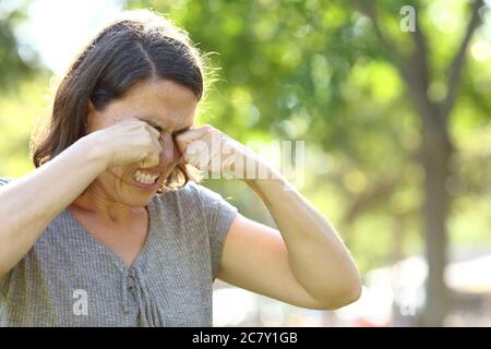 Donna di mezza età che gratta occhi prurito in piedi in un parco in estate Foto Stock