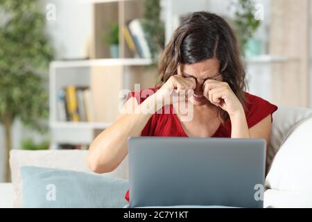 Donna adulta con un computer portatile che graffia gli occhi pruriginosi seduti nel divano nel soggiorno di casa Foto Stock