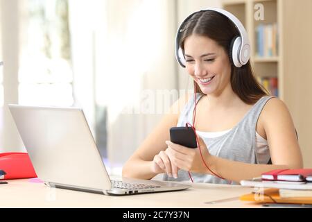 Una studentessa felice con un computer portatile ascolta la musica sulle cuffie e sullo smartphone seduto su una scrivania a casa Foto Stock