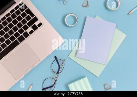 Flaylay di computer portatile, occhiali e cancelleria su uno sfondo blu pastello. Concetto di spazio di lavoro con forniture per ufficio. Foto Stock