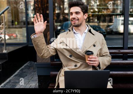 Giovane attraente uomo sorridente in trench cappotto con laptop e caffè gioiosamente sventolando la mano ciao sulla strada Foto Stock
