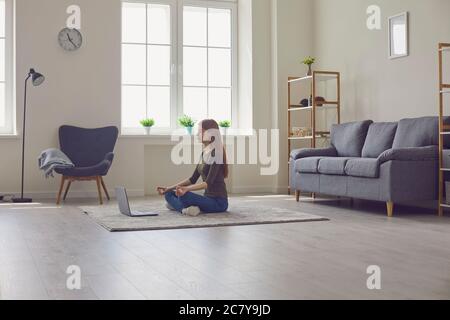 Meditazione yoga online. Donna con computer portatile seduto in Lotus posa sul tappeto in casa Foto Stock