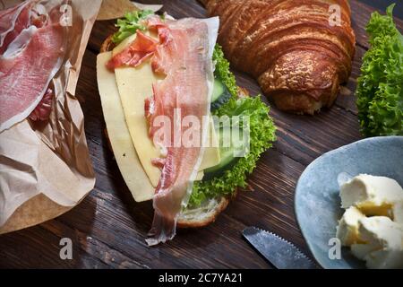 panino di croissant con fette di carne di jamon, foglie di lattuga verde, cetrioli freschi, formaggio, burro e coltello, su tavola rustica in legno, piatto Foto Stock