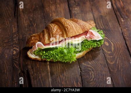 un panino di croissant con fette di carne di jamon, foglie di insalata di lattuga verde, cetrioli freschi e formaggio, su una rustica superficie di tavola in legno, copyspace Foto Stock