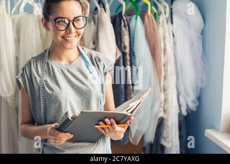 Giovane donna dressmaker scegliendo materiale dal catalogo in studio. Adattare l'aspetto attraverso i tessuti mentre si è in piedi in un laboratorio di cucitura Foto Stock