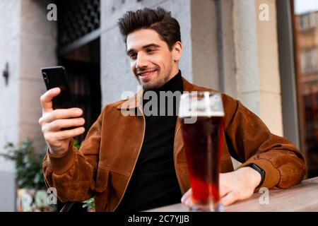 Giovane attraente uomo informale felicemente utilizzando il cellulare mentre beve birra in un caffè sulla strada Foto Stock