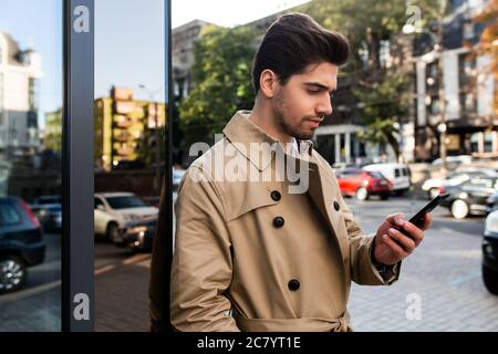 Vista laterale di un giovane e attraente uomo d'affari in trench cappotto utilizzando il cellulare sulla strada Foto Stock