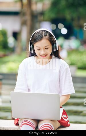 Sorridente ragazza asiatica adolescente in cuffie seduto in parco e lavorando su computer portatile o programmazione Foto Stock