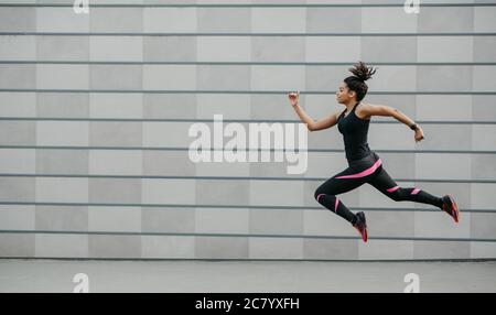 Eccellente formazione in esecuzione. Ragazza afroamericana in abbigliamento sportivo si è congelata in aria sopra terra, saltando Foto Stock