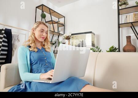 Bella donna bionda sorridente giovane seduta sul divano e lavorando sul computer portatile quando si è a casa a causa della quarantena Foto Stock