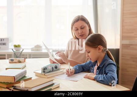 Ritratto con vista laterale dai toni caldi della bambina cute che scrive prova mentre studia a casa con la madre o il tutor che la aiuta, copia spazio Foto Stock