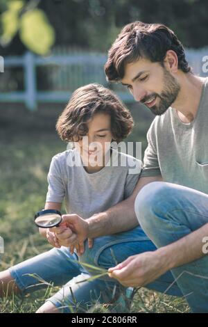 Ragazzo dai capelli scuri e suo padre che guarda la pianta attraverso la lente d'ingrandimento e che guarda interessato Foto Stock