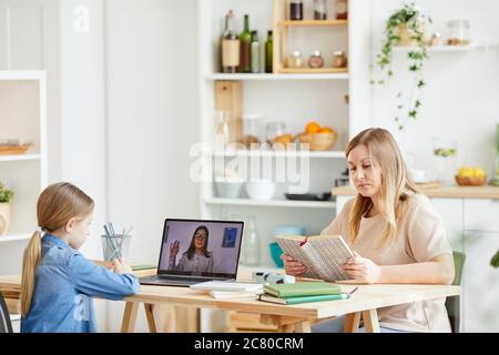 Ritratto laterale di bambina che usa il computer portatile durante la lezione online con tutor o insegnante mentre si siede alla scrivania in accogliente cucina interna con madre wa Foto Stock