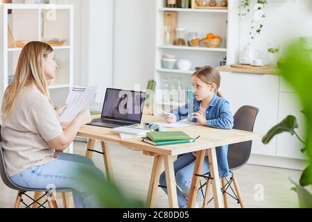 Ritratto laterale di madre che fa i compiti con la figlia carina mentre si siede alla scrivania e guardando i video della scuola online, spazio di copia Foto Stock