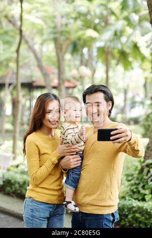 Ridendo giovani genitori e il loro figlio ridendo quando si prende selfie insieme in parco Foto Stock