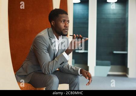 Ritratto di bell'uomo d'affari afro-americano che registra il messaggio vocale tramite smartphone mentre si siede nella zona lounge dell'ufficio moderno o coworking sp Foto Stock