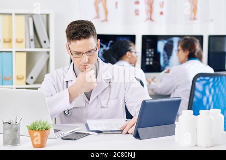 Serio medico accigliato che controlla il documento con le statistiche di incidenza quando sedendo al suo tavolo Foto Stock