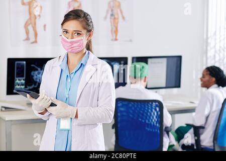 Giovane dottore in guanti di gomma e maschera medica in piedi con il computer tablet in mani, i suoi colleghi discutere di dati di ricerca su computer scre Foto Stock