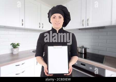 giovane cuoca donna in uniforme nera che mostra gli appunti vuoti nella cucina moderna Foto Stock