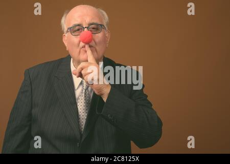 Uomo d'affari anziano in sovrappeso che indossa il naso di clown su sfondo marrone Foto Stock