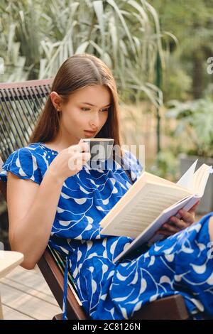 Giovane attraente donna con capelli marroni in abito blu bere caffè e leggere attentamente libro su sedia in legno ponte nel parco Foto Stock