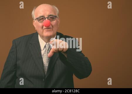 Uomo d'affari anziano in sovrappeso che indossa il naso di clown su sfondo marrone Foto Stock