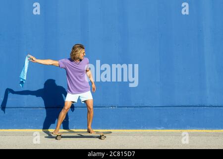 Giovane biondo con sciarpa blu che va a skate con fondo blu. Estate, concetto di surf. Foto Stock