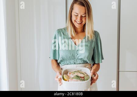 Giovane donna che tiene vassoio con pollo ed erbe aromatiche e. sorridente Foto Stock