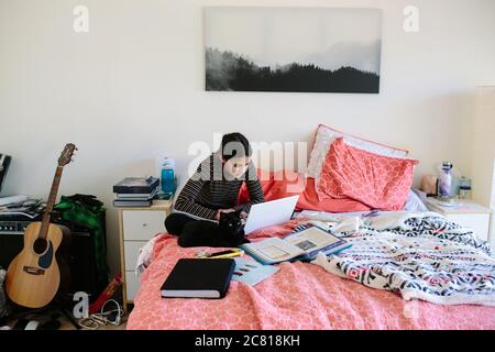 La ragazza della scuola media si siede con il suo gatto mentre imparando in linea sul suo letto Foto Stock