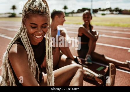 Atleta sorridente seduto in pista con altri corridori che si allungano in background. Sportivo che si rilassa e si allunga dopo una corsa pratica sulla t Foto Stock