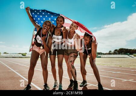 Ritratto di giovane squadra di atlete che godono la vittoria. Gruppo diversificato di runner con medaglie che celebrano il successo tenendo una bandiera degli Stati Uniti sull'ippodromo. Foto Stock