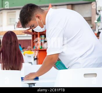 Punta Umbria, Huelva, Spagna - 10 luglio 2020: Un cameriere in maschera facciale pulisce un tavolo della barra con liquido disinfettante Foto Stock