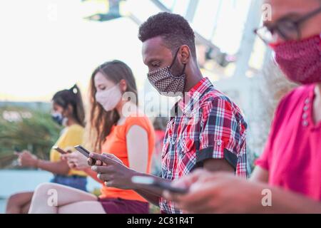 Amici che usano il loro smartphone in covid 19 volte protetto con maschera facciale - giovani persone che usano il dispositivo mobile a distanza all'aperto